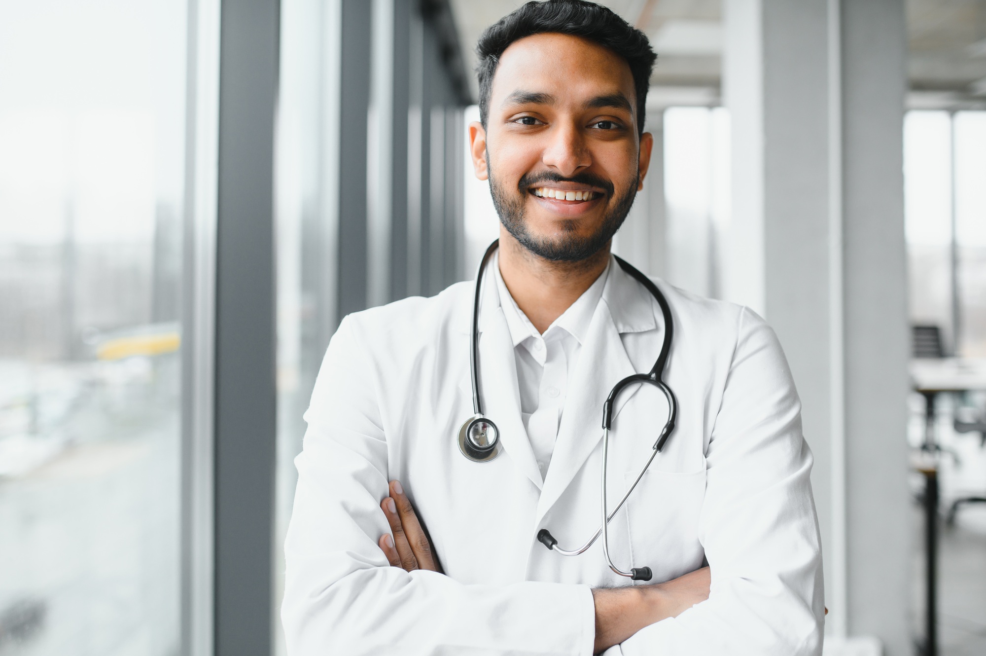 Portrait of male indian doctor wearing white coat having open door on clinic corridor as background